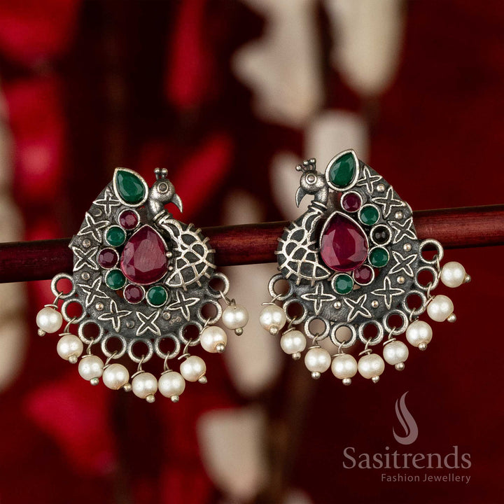 Traditional oxidised silver earrings with intricate peacock motif and ruby-green waterdrop stones framed in floral design – ideal for vibrant festive jewellery styling – Sasitrends