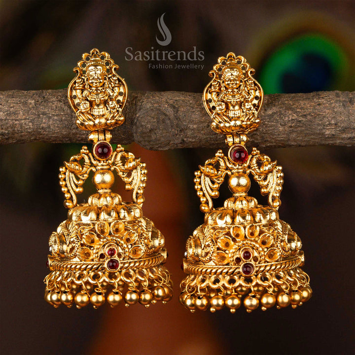 Ornate temple-style matte gold jhumka earrings crafted with ruby stone highlights, sculpted Lakshmi artwork and elegant peacock carvings – ideal for showcasing festive ethnic charm – Sasitrends