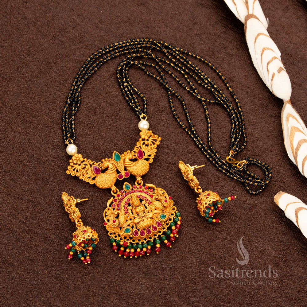 Exquisite matte gold temple necklace jewellery set featuring goddess Lakshmi and regal peacock motifs, paired with ornate ruby-green stone jhumkas—crafted in a heritage bridal design inspired by South Indian tradition - Sasitrends