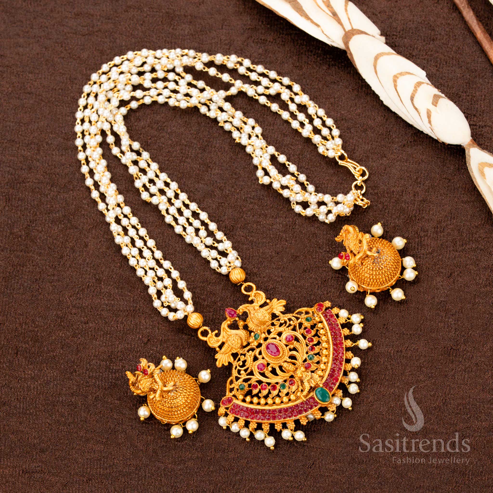 Matte gold temple jewellery set with ruby-green stone highlights, a detailed Lakshmi pendant, peacock artistry, and pearl hangings—an antique bridal grandeur piece designed for luxurious ethnic styling and bridal charm - Sasitrends
