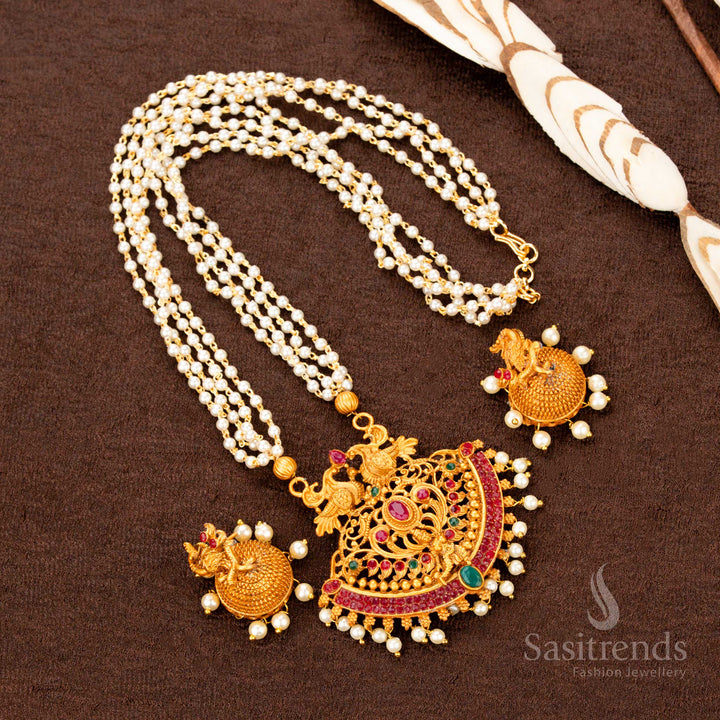 Matte gold temple jewellery set with ruby-green stone highlights, a detailed Lakshmi pendant, peacock artistry, and pearl hangings—an antique bridal grandeur piece designed for luxurious ethnic styling and bridal charm - Sasitrends